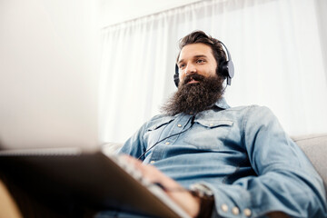 Bearded entrepreneur using laptop and headphones at home.