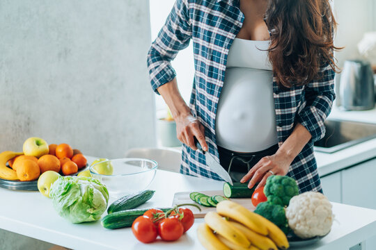 Unrecognisable pregnan woman cook salad cutting cucumber on wooden cutting board putting fresh vegetables and fruit on table in kitchen. Pregnancy, maternity, healthy lifestyle concept. Copy space.