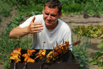 Young man cooking at a barbecue grill chicken wings. Man smelling and enjoying picnic food....