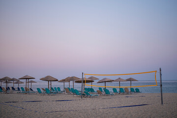 Beach Volleyball net on sandy beach early in the morning, with umbrellas and sunbeds, sea and blue sky in background.