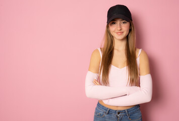 Beautiful girl wearing casual clothing and black cap posing with arms folded isolated over pink background.