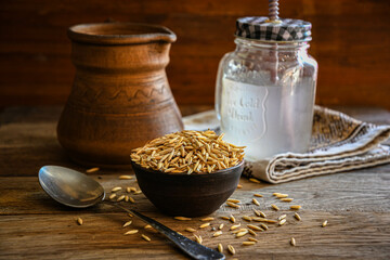On the wooden kitchen table are a ceramic bowl of oat grains, a glass cup with oat kvass on a kitchen napkin, and an earthen jug. Village life. - obrazy, fototapety, plakaty