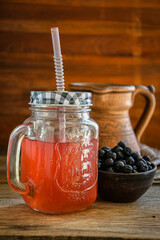 On the wooden kitchen table, there is a ceramic bowl with black rowan berries, a glass cup with black rowan kvass on a kitchen napkin, and an earthen jug. Village life. Healthy drinks. Ethnoscience.