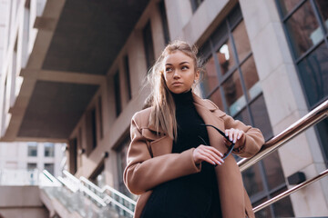 A young beautiful girl in a beige coat with sunglasses stands on the street