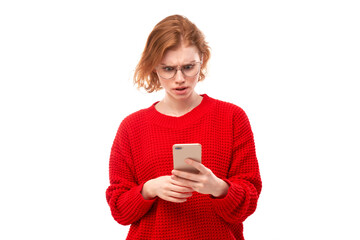 Pensive thinking redhead girl looks into smartphone screen with upset face in glasses and red clothes isolated on white studio background