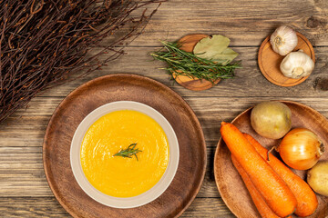 Carrot and potato soup bowl on a drift wood table with vegetables in a wood plate, dry wood branches, garlic, laurel and rosemary. Flat lay.