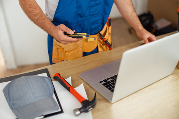Fototapeta na wymiar Cropped shot of repairman worker in uniform wearing tool belt, holding utitlity knife and using laptop while standing indoors