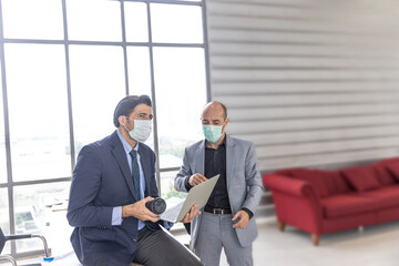 Two male managers with hygienic face masks are using computer to discuss business project in the office as a new normal practice during COVID-19 pandemic. New business model to prevent COVID-19