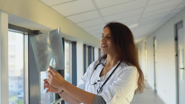 Female indian doctor or intern examines x-ray of lungs, holding it in hands indoors. Specialist holds transparent image of chest in arms, and carefully researches it