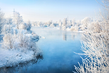 Trees on the bank of a blue river in white snow frost in the Moscow region on a winter sunny evening