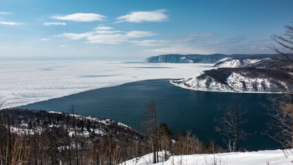 A non-freezing river flows out of a frozen lake. White ice and blue water. A mountain range on the background of an azure sky with clouds. Wooded and snow-covered shores. Baikal and Angara