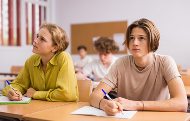 Teenage boy and girl sitting at table in classroom and working on tasks during lesson. Teacher standing near and looking at pupil's copybook.