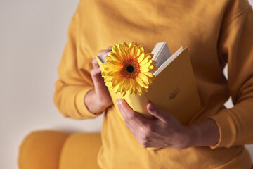 A yellow book with a flower bookmarked in the hands of a woman on a yellow sweater background. Front view.