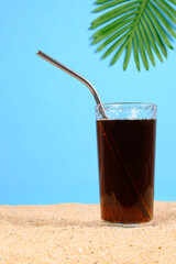 Cold drink in glass on sand against blue background with palm leaf