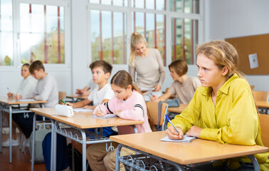 Focused school girl sitting at school desk in classroom on background with classmates