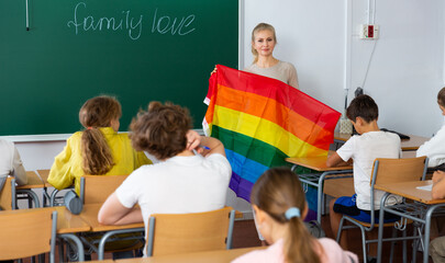 Female teacher holding rainbow-colored LGBT community flag and explaining it meaning for kids...