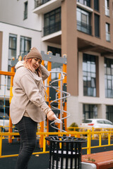 Vertical shot of overweight woman in warm hat and jacket using antibacterial wet wipes to clean disinfect hands and throws them in trash, standing on background on modern building in cloudy autumn day