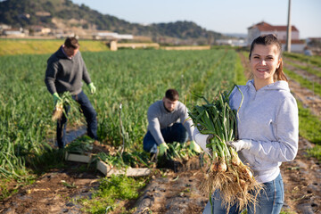 Portrait of cheerful young woman engaged in green onions harvesting on family farm plantation