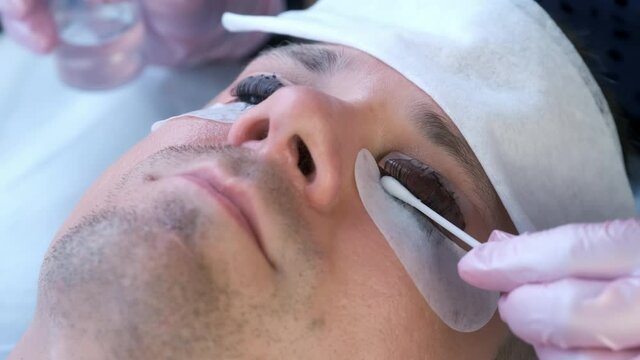 Beautician is wiping black paint from man's eyelashes on laminating lash procedure. Cosmetologist is making lash lifting lamination in beauty salon, cosmetology clinic, closeup face of client.