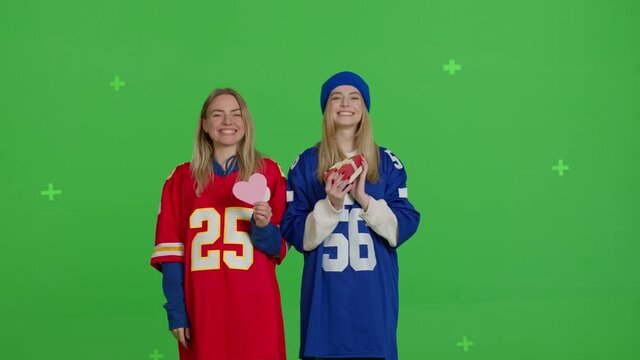 Two girlfriends in football t-shirts holding gifts and heart-shaped card. Friendship love and valentine's day. Young caucasian women having fun over green screen background. Chroma key