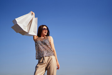 Attractive brunette glam girl in sunglasses with shopping bags standing in bright clothes at sunny day. Caucasian woman with shopping bags against blue sky background. High quality photo