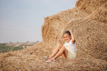 Naklejka na ściany i meble Portrait of young Caucasian little girl having fun sitting on haystacks throwing hay at someone with happy playful face. Time away from city in country field with tons of hayricks.