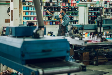 Male worker mixing colors for screen printing in a workshop and using a smartphone