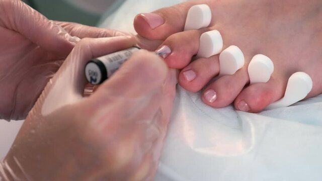 Pedicurist master is applying second coat of shiny soft pink gel shellac on woman's toes in beauty clinic, closeup view. Hygiene and care for feet. Professional pedicure in cosmetology clinic.