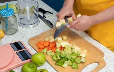 Women's hands cut an apple with a sharp knife on a wooden board, where sliced vegetables and fruits lie. The process of making smoothies. Selective focus.
