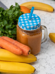 Freshly prepared smoothie in jar with straw. There are carrots, bananas, and greens nearby. Close-up from above. Selective focus.