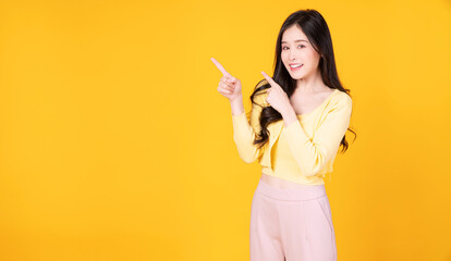 Cheerful Asian woman use finger pointing to product or empty copy space standing over isolated on yellow background. Surprised model young girl present content. Advertisement presenting concept.