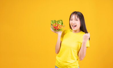 Excited cheerful Asian woman slim body holding glass bowl with organic fresh vegetables raised own arm for good health while standing over isolated yellow background. Health care food concept.