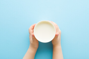 Toddler hands holding white plastic glass of fresh milk on light blue table background. Pastel...