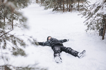 Fototapeta na wymiar Happy teenager boy lying on snow in winter forest. Child having fun outdoors. Joyful adolescent playing in snow at snowfall. Laughing smiling kid walking in winter park in cold weather