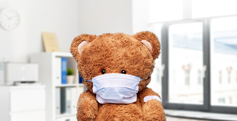 vaccination, healthcare and pandemic concept - teddy bear toy in protective mask with patch on paw over medical office at hospital background