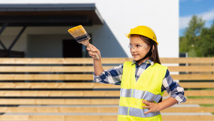 building, construction and profession concept - smiling little girl in protective helmet and safety vest with paint brush over house background