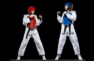 Dynamic portrait of two young women, taekwondo athletes training together isolated over dark...