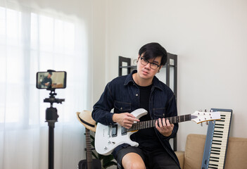 Obraz premium Young asian musician teaching the rock guitar class online live streaming on smartphone with student. Korean man playing electric guitar singing a song and record by cell phone shaed online social