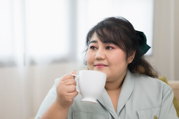 Happy young women's plus sitting on sofa drinking coffee in living room at home. Body positive plus size female lives happily and is proud of herself.