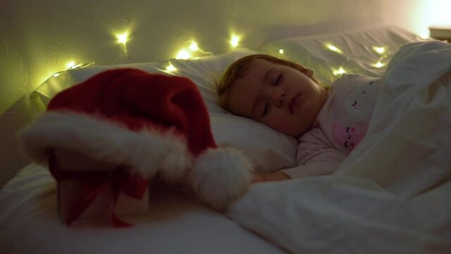 Cute Chuby Little Toddler Baby Girl 1-2 Years Old Sleeping In Pink Pajama On White Bed In Dark Bedroom. Child Have Relax Rest Time At Night Garland on Pillow. Childhood, Christmas, Parenthhood Concept
