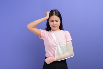 Fototapeta premium Portrait of young woman stressed and frustrated with an injured arm in a sling over blue background in studio, insurance and healthcare concept