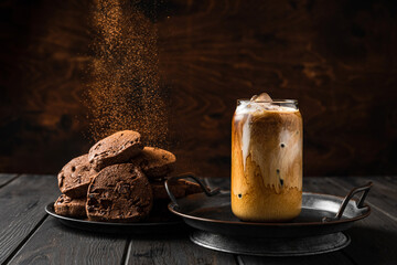 A glass of iced coffee with milk and chocolate cookies on a plate, cocoa powder falling