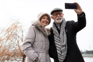 An elderly couple walks around the waterfront in an urban area and takes a selfie. The couple is smiling and looking happy in love.