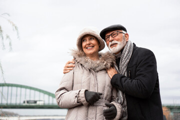 An elderly couple is walking around the waterfront in an urban area. The couple is smiling and looking happy in love.