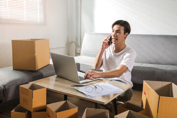 Cheerful Asian male freelance sits on floor at home and works on a laptop computer having a new customer order on a cellphone with the postal parcel for shipping. Succeed Online selling concept ideas