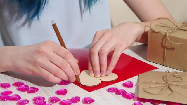 Valentines Day craft DIY. A female hand draws a contour of a heart shape on red paper on a table with gifts and decorations