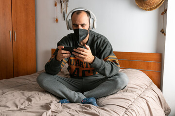 Man with headphones, mobile phone and face mask, sitting on his bed and confined to his room