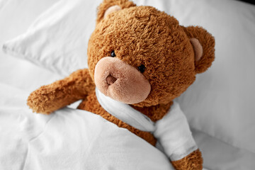 medicine, healthcare and childhood concept - ill teddy bear toy with bandaged paw lying in bed