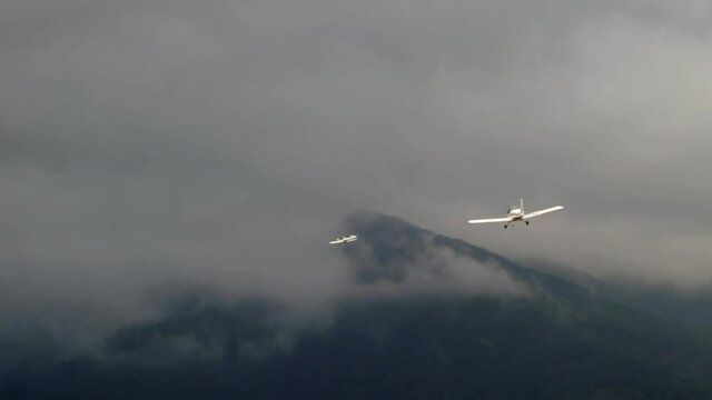 Two small airplanes flying towards mountains of a cloudy day