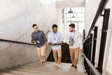 business, people and corporate concept - group of men with conference badges, folder, tablet pc computer and smartphone walking up office stairs